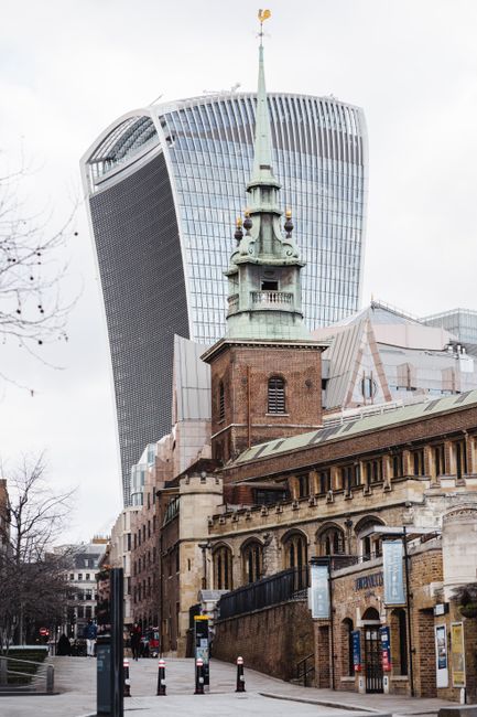 A view of a historic brick church with a tall, ornate spire topped by a weather vane, situated on a city street. In the background, the modern, glass-clad 20 Fenchurch Street skyscraper, also known as the Walkie Talkie, features a distinctive curved design and reflective windows. The street scene includes several pedestrians, a black metal fence, and pavement with bollards and signage. The image depicts a contrasting blend of historic architecture and contemporary commercial building, illustrating the urban environment where house removals and furniture transport services might take place, especially in the context of moving logistics near iconic London landmarks, as described on Bowmanandvan.org.uk.