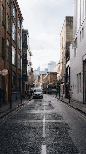 A narrow urban street in Bow, London, with tall multi-story buildings on both sides, featuring a mix of brick and modern facades. The street is currently used for vehicle and foot traffic, with a white van parked near the curb on the left and several pedestrians walking along the pavement. In the foreground, there is a section of the wet asphalt road with a central dashed white line, indicating recent rain or cleaning. The street appears to be part of a busy area suitable for home relocation and furniture transport services, with Bow Man and Van occasionally involved in loading or unloading furniture and boxes from nearby properties. Visible in the background are taller city buildings under a cloudy sky, emphasizing the urban environment. The scene reflects typical conditions for house removals, with a focus on the street layout, building styles, and possible vehicle manoeuvres during packing and moving processes in a dense city environment.