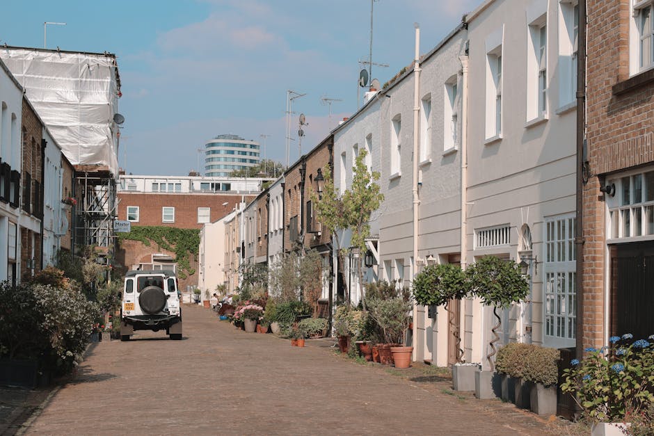 A narrow, cobbled residential street with closely spaced terraced houses on both sides, featuring a mixture of brick and white-painted exteriors. Several potted plants and small trees are positioned along the pavement in front of the properties. A white van, belonging to Bow Man and Van, is parked on the street near the curb, ready for a home relocation or furniture transport. The van is adjacent to the property’s doorway, with some items possibly prepared for loading. Scaffolding is visible on one building indicating ongoing work, while television antennas and satellite dishes are mounted on rooftops beneath a partly cloudy sky. The scene depicts the logistical challenges of moving goods through a tight street, incorporating packing materials such as cardboard boxes and blankets used to protect furniture during the loading process. Overall, the image captures the environment typical for a professional removals service operating within urban streets with limited parking and tight access.