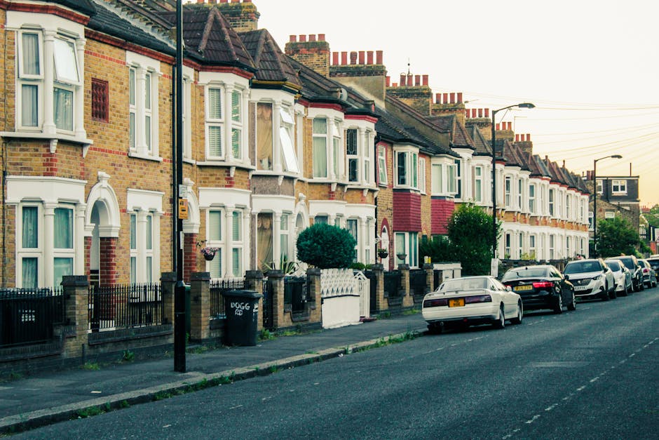 A narrow urban street in Bow, London, with tall multi-story buildings on both sides, featuring a mix of brick and modern facades. The street is currently used for vehicle and foot traffic, with a white van parked near the curb on the left and several pedestrians walking along the pavement. In the foreground, there is a section of the wet asphalt road with a central dashed white line, indicating recent rain or cleaning. The street appears to be part of a busy area suitable for home relocation and furniture transport services, with Bow Man and Van occasionally involved in loading or unloading furniture and boxes from nearby properties. Visible in the background are taller city buildings under a cloudy sky, emphasizing the urban environment. The scene reflects typical conditions for house removals, with a focus on the street layout, building styles, and possible vehicle manoeuvres during packing and moving processes in a dense city environment.