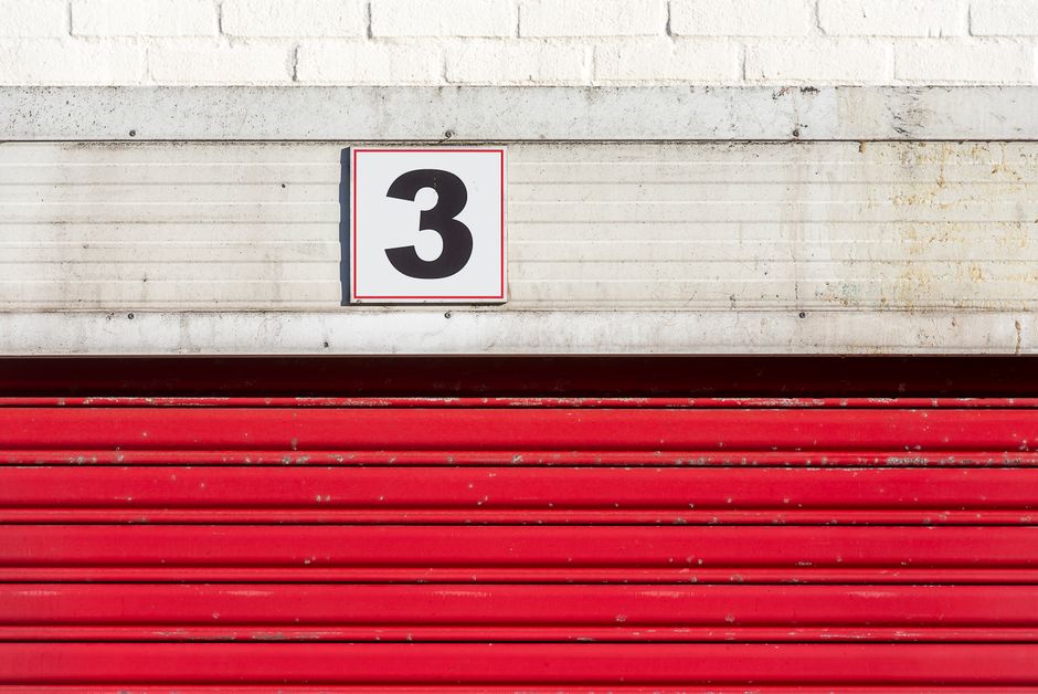 A metal roller shutter door painted in red, with visible horizontal ridges, is partially closed over a white brick building. Above the door, there is a white rectangular sign with a black number '3' i